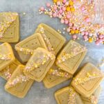 Several rectangular River Road Vegetarian Citrus Shortbread cookies topped with a clear glaze and a diagonal sprinkle pattern in pink, yellow, and white, with extra nonpareil sprinkles scattered nearby.