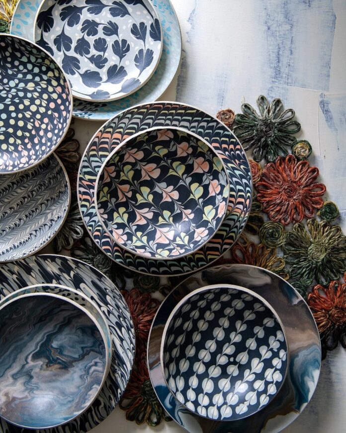 Arrangement of marbled and patterned ceramic plates and bowls by Morgan Levine, featuring slipware designs like flowing blue swirls and leaf motifs in dark blue, white, and pale yellow, resting on colorful woven mats.