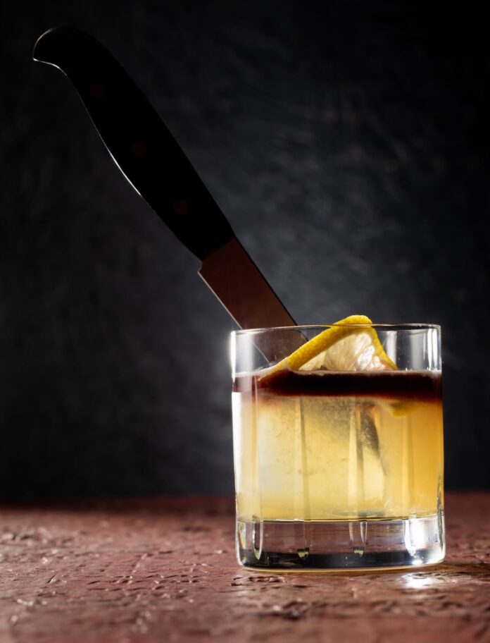 A cocktail for benoit blanc in knives out with a in a glass in a pale yellow color with a red float on top, lemon twist and knife through it all.