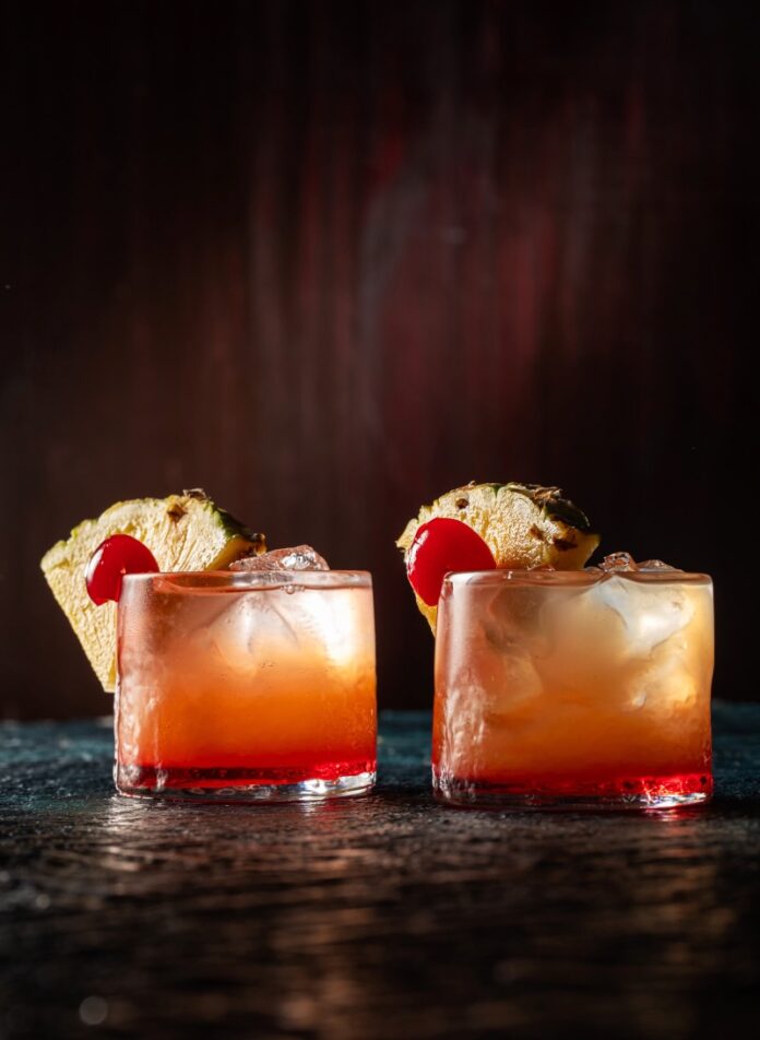 Two glasses of an orange cocktail with cherry and pineapple wedge garnishes.