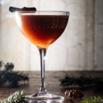 A coupe glass with a brown Three Wisest Men cocktail and a toasted cinnamon stick garnish as sprigs of pine trees lay on the table.