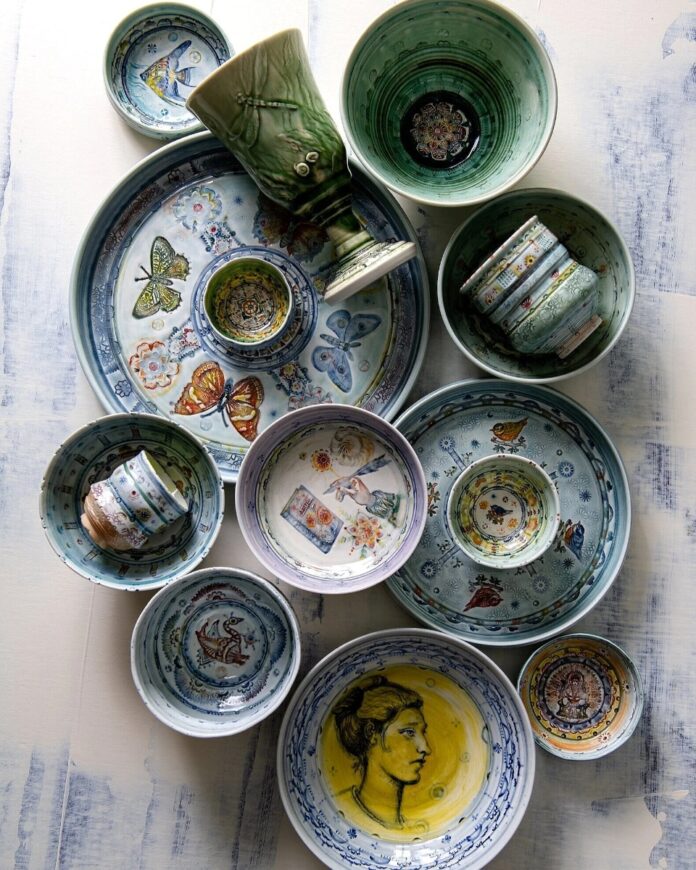 Arrangement of artisan ceramic bowls and platters by Janis Cutler Gear, featuring various painted designs, including butterflies, a classical portrait, birds, flowers, and intricate patterns in shades of blue, green, and yellow.