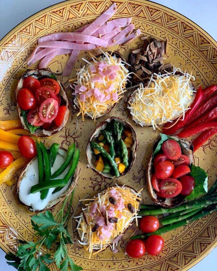 A Flotilla of Potato Boats TABLE Magazine Potato boat halves on a yellow patterned platter, filled with various toppings like cheese, ham, tomatoes, asparagus, bell peppers, sour cream, and herbs, ready for serving.