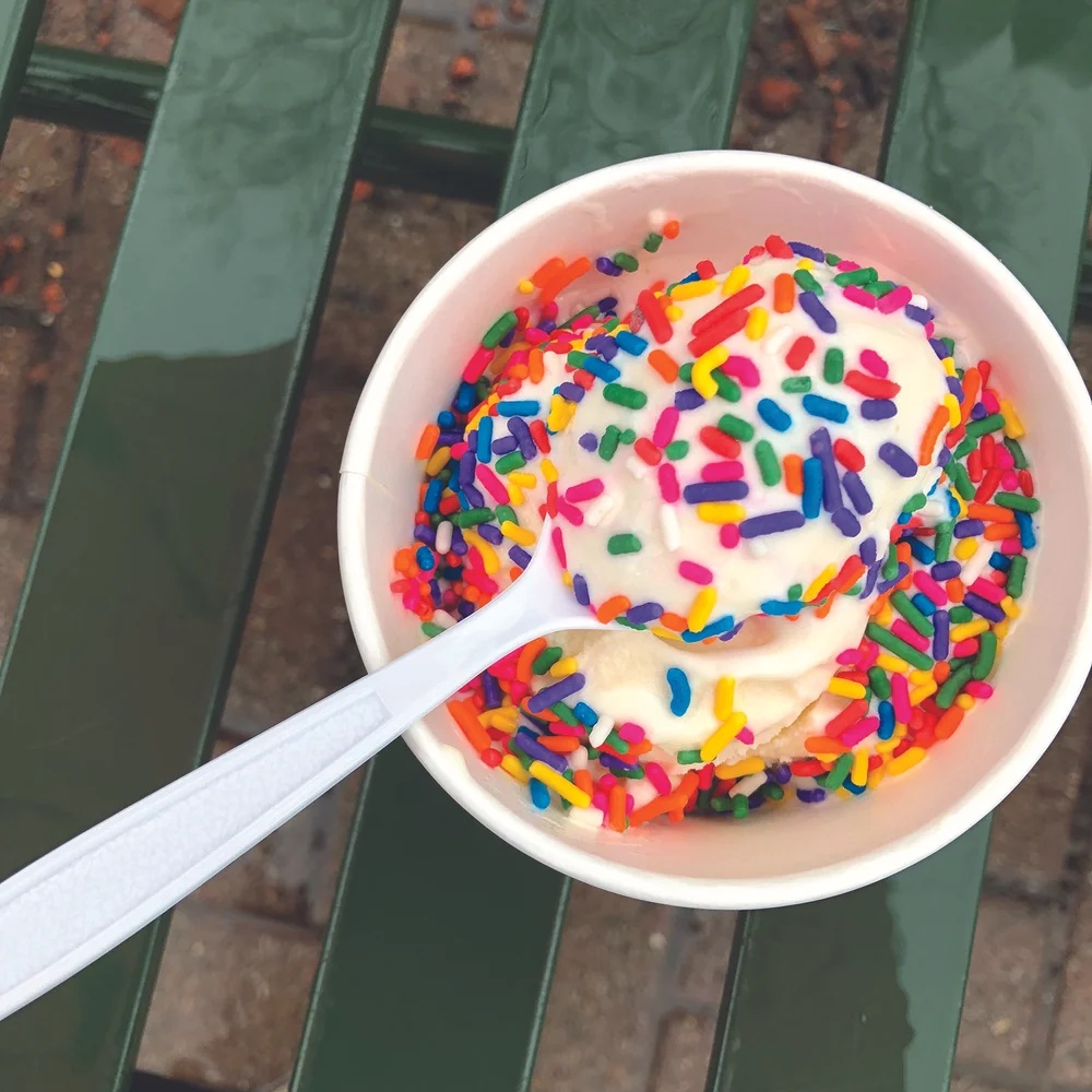 A small white cup of vanilla custard or ice cream covered in colorful rainbow sprinkles, with a white plastic spoon resting on top, set on a green metal bench.