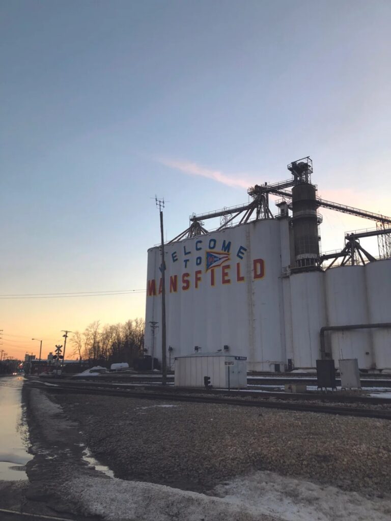 A large, white industrial grain silo with the painted words "WELCOME TO MANSFIELD" in front of railroad tracks and a small patch of snow at sunset in Ohio.