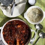 Dark stew of lamb shank, beans, and carrots, served next to a bowl of white rice, silverware, and a small bowl of salt on a bright.