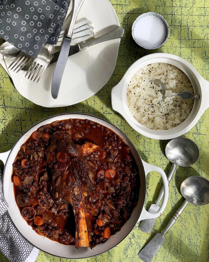 Dark stew of lamb shank, beans, and carrots, served next to a bowl of white rice, silverware, and a small bowl of salt on a bright.