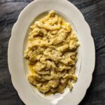 A white oval platter piled high with Gnocchi Sbatúi, or beaten gnocchi, covered in melted butter, cheese, and pepper, set on a dark, gray marble surface.