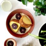 Two slices of moist, golden poundcake served in a rustic brown bowl, covered in a white drizzle or custard and garnished with dark red blood orange slices, with a glass of milk and houseplants nearby.