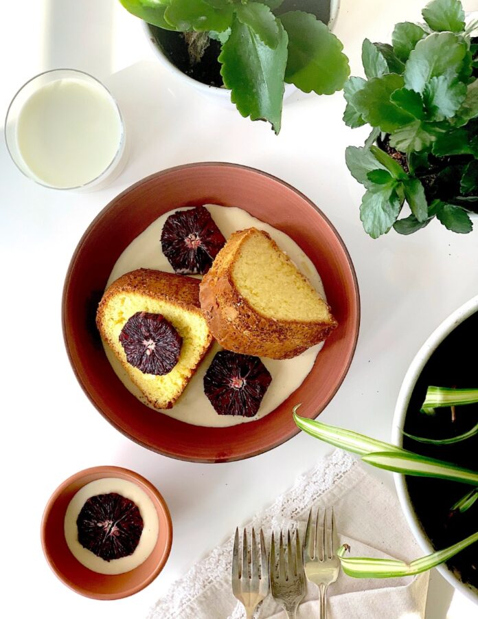 Two slices of moist, golden poundcake served in a rustic brown bowl, covered in a white drizzle or custard and garnished with dark red blood orange slices, with a glass of milk and houseplants nearby.