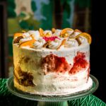 A cake with white icing decorated with orange slices and cranberry displayed on a glass cake plate on a table with a green table cloth.