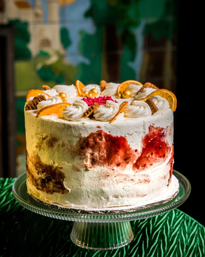 A cake with white icing decorated with orange slices and cranberry displayed on a glass cake plate on a table with a green table cloth.