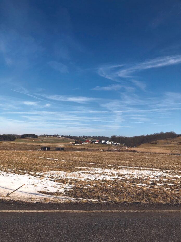A rural Ohio field in winter with patches of snow on brown grass, rolling hills in the distance, and farm buildings under a bright blue sky with wispy clouds.