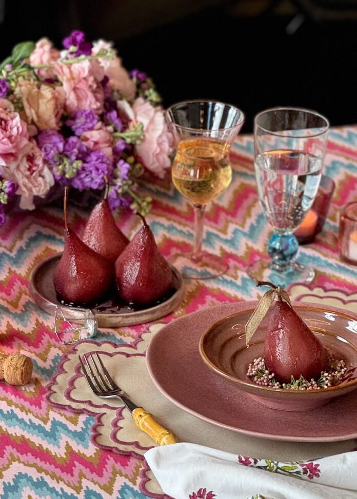 Three poached pears in a red color sit on a small plate while a larger plate and bowl holds another full pear off to the side of the table setting.