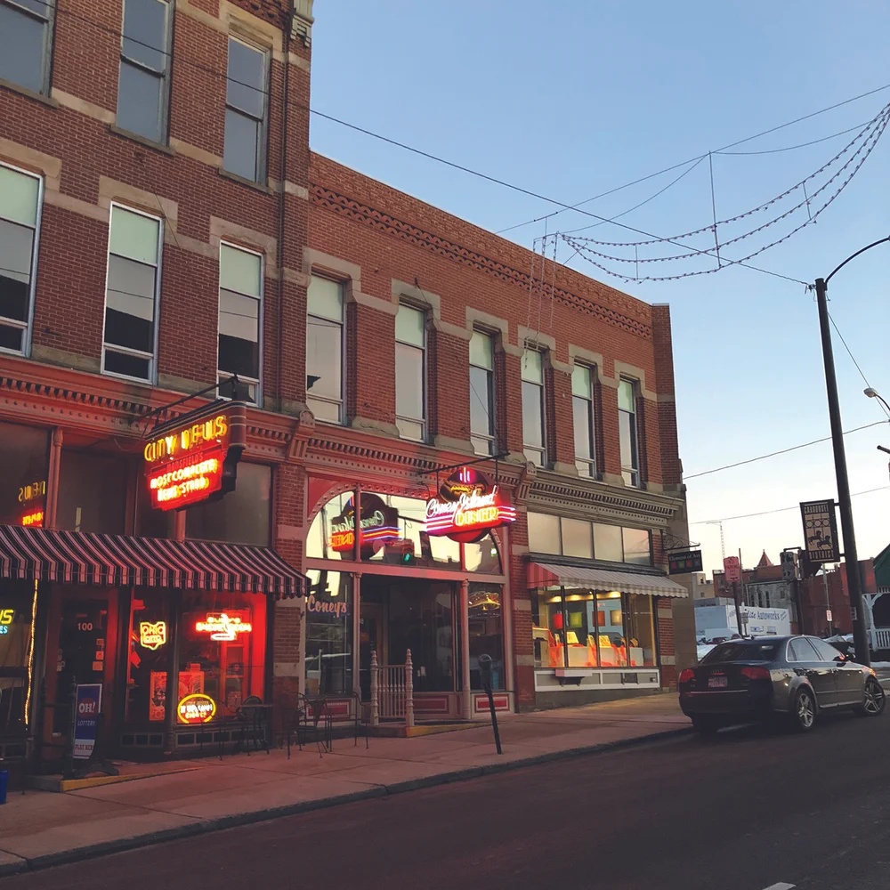 A sunset street view of red brick buildings in an Ohio downtown, featuring bright red neon signs for 'CITY NEWS' and 'SCHLEPP'S RESTAURANT' on the ground floor, with a car parked on the street.