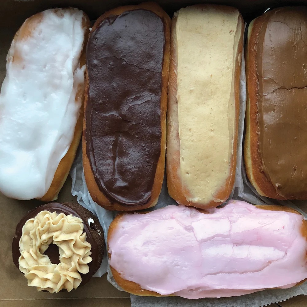 Six variously glazed donuts, including white, chocolate, pink, and butterscotch/maple long johns, and one chocolate ring donut topped with beige piped frosting.