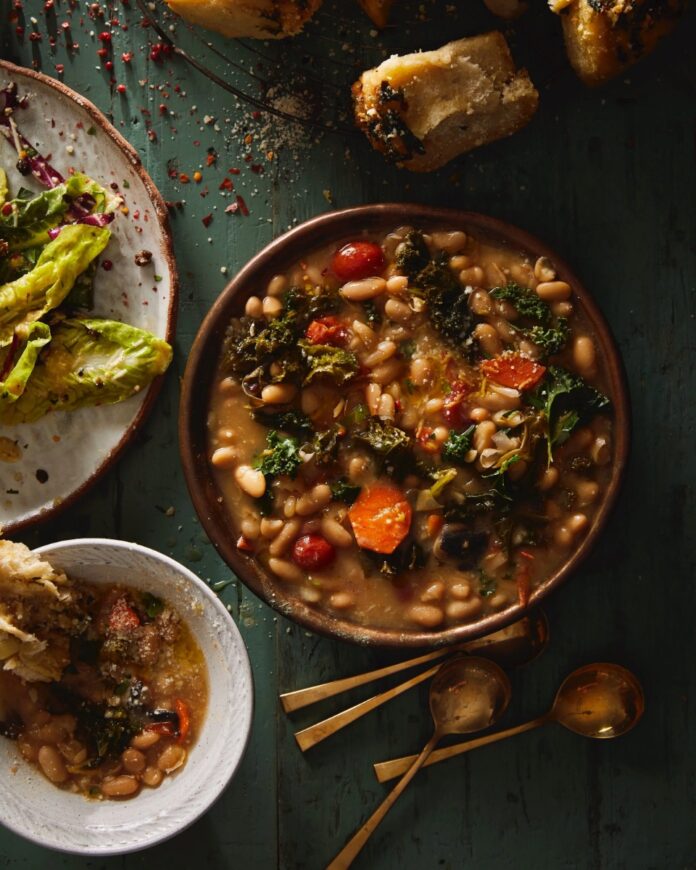 A large bowl of steaming white beans and chopped kale stew garnished with carrots and tomatoes, served with crusty bread/garlic knots and a side salad on a green wooden table.