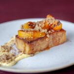 A plated dessert featuring a thick slice of golden-brown bread pudding topped with caramelized peach slices and crumble, resting on a creamy butterscotch sauce on a white plate.