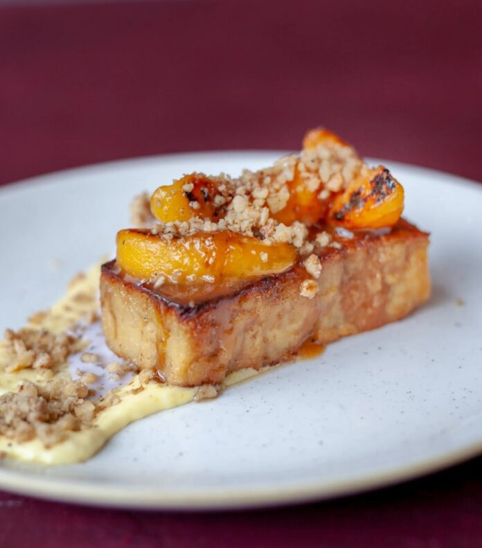 A plated dessert featuring a thick slice of golden-brown bread pudding topped with caramelized peach slices and crumble, resting on a creamy butterscotch sauce on a white plate.