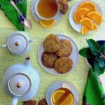 Golden-brown cookies served with cups of tea and slices of fresh orange on a light yellow textured surface, next to a white teapot and a green floral napkin.