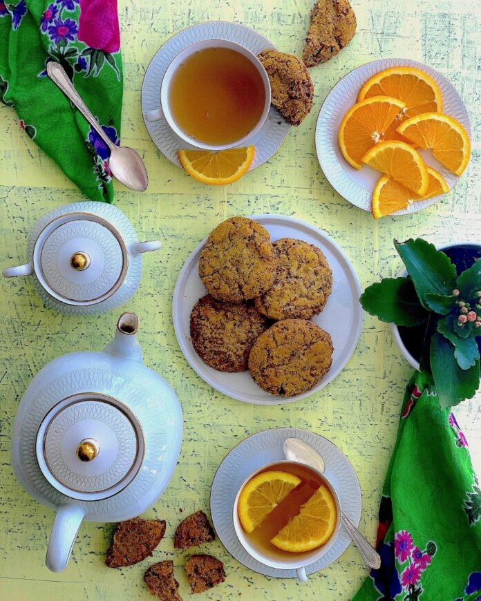 Sunshine Cookies TABLE Magazine Golden-brown cookies served with cups of tea and slices of fresh orange on a light yellow textured surface, next to a white teapot and a green floral napkin.