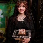 A women dressing in black showing the camera a large vegan brownie decorated with peppermint candies on a glass cake pedestal.