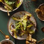 A econstructed Caesar salad with whole gem lettuce leaves, red radicchio, and creamy dressing on a gray plate, served next to two bowls of soup/stew and chunks of bread on a green wooden table.