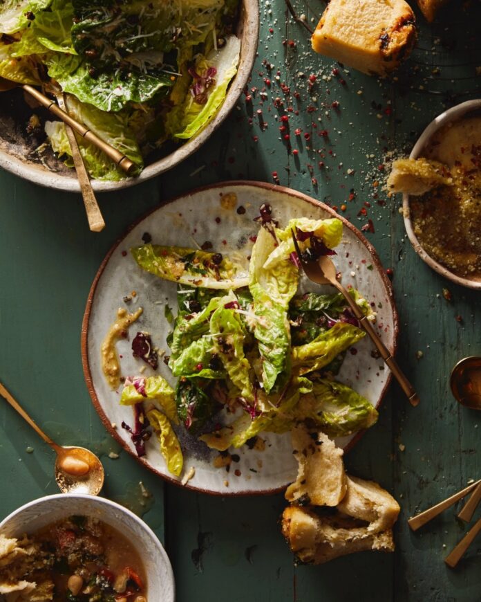 A econstructed Caesar salad with whole gem lettuce leaves, red radicchio, and creamy dressing on a gray plate, served next to two bowls of soup/stew and chunks of bread on a green wooden table.