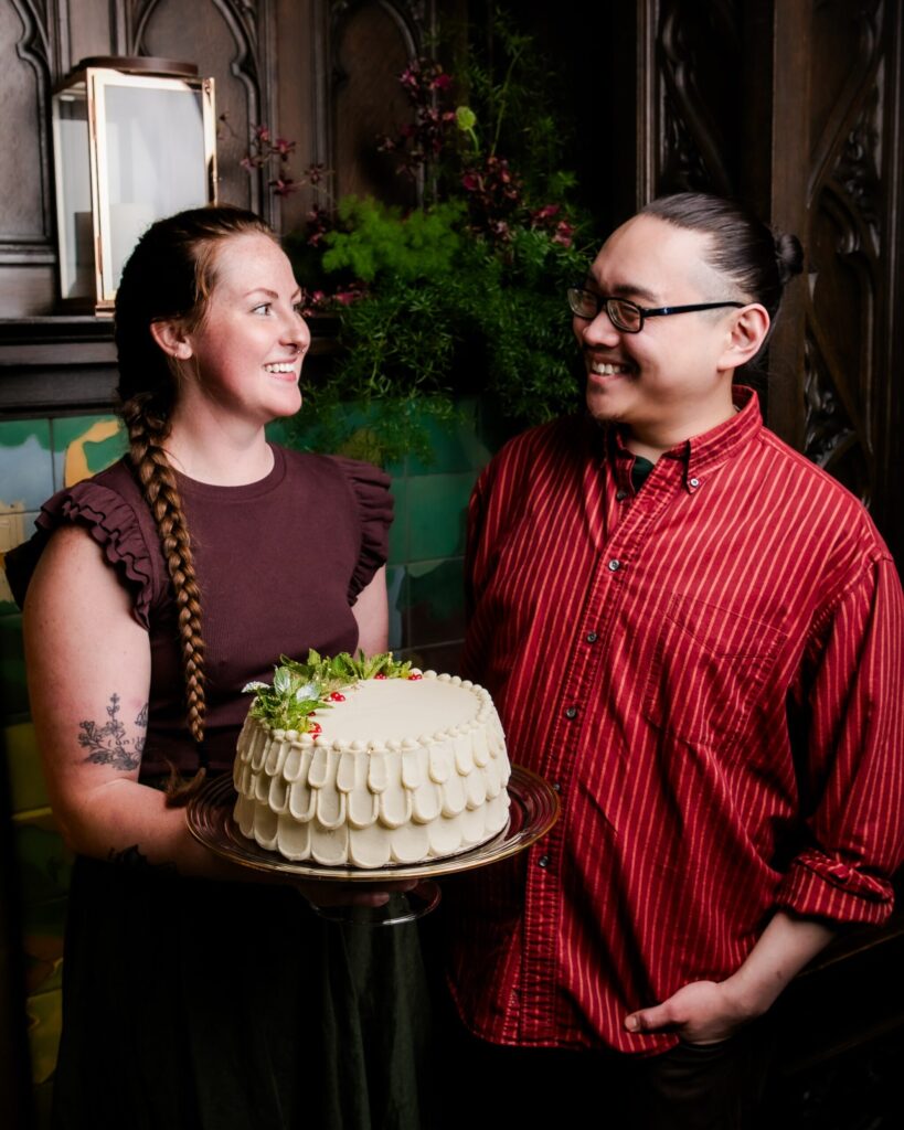 A man and a women standing together while the woman displays her white iced cake for the camera.