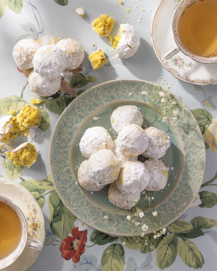 Bridgerton Teatime Pecan Balls TABLE Magazine A green plate full of Pecan Balls stacked in a tower as some sit open with yellow cake on the table.
