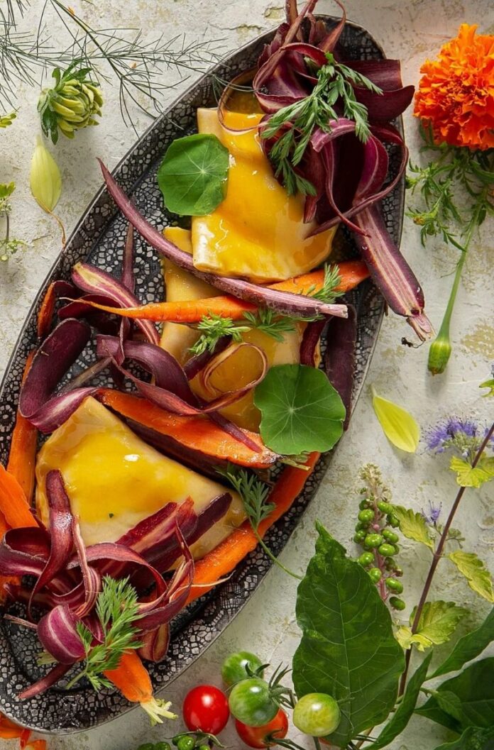 A plate of carrot pierogies garnished with carrot ribbons and fresh herbs, surrounded by flowers and green and red vegetables.