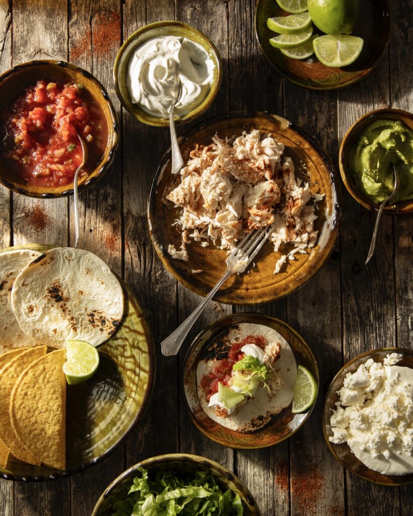 Various plates full of taco toppings like chicken, salsa, sour cream, lime juice, and more.