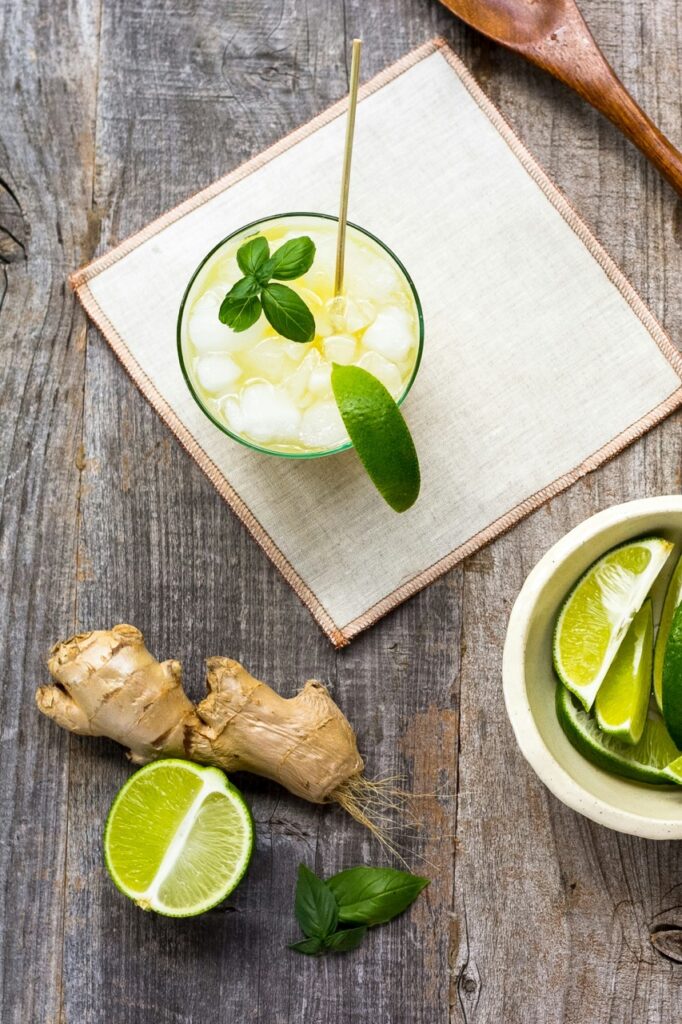 A glass of lemonade with basil, ginger, and line sits on a white square on a wood table.