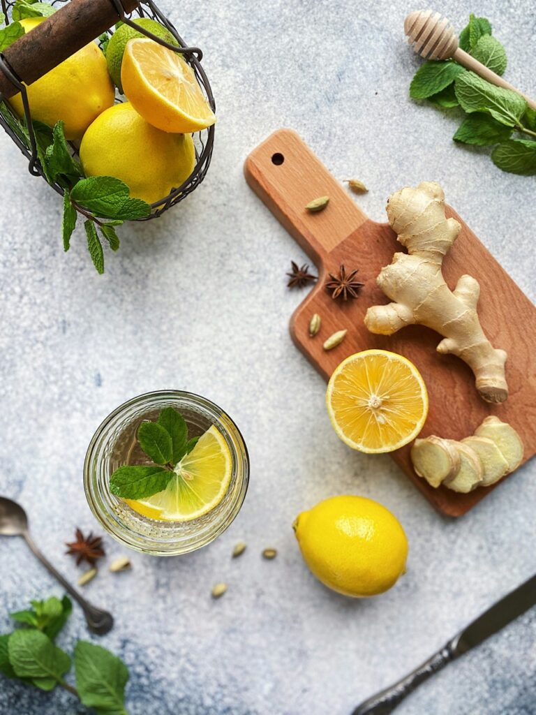 Ginger and lemon slices sit on a cutting board beside a bowl of lemons and a glass of water with a slice of lemon in it. 