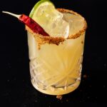 A Hot Bunny Smash in a yellow color sits in a cocktail glass with a tajin rim, lime slice, and hot pepper garnish.