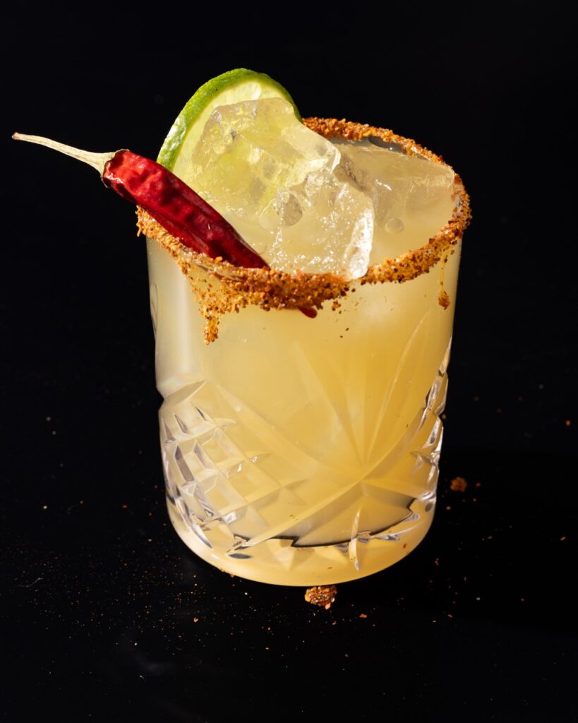 A Hot Bunny Smash in a yellow color sits in a cocktail glass with a tajin rim, lime slice, and hot pepper garnish.