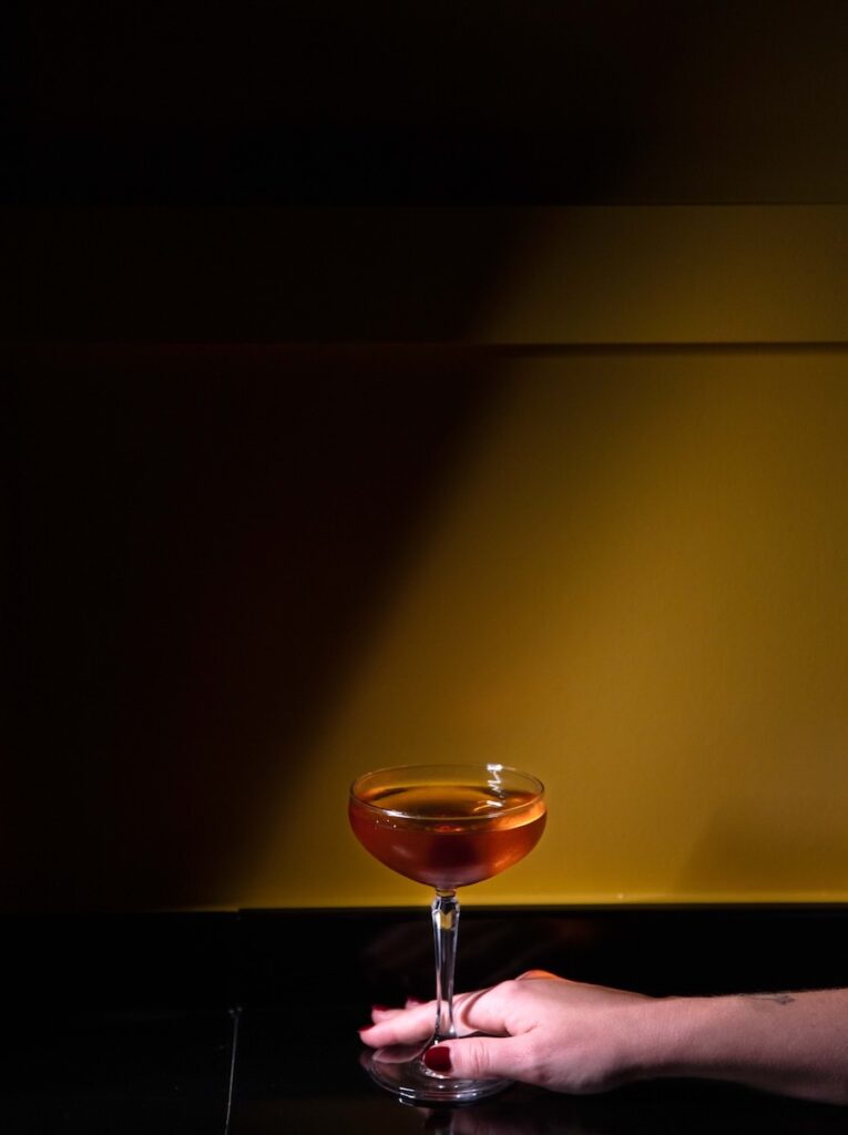 An amber cocktail sits against a dimly lit wall as a woman's hand pushes it forward.