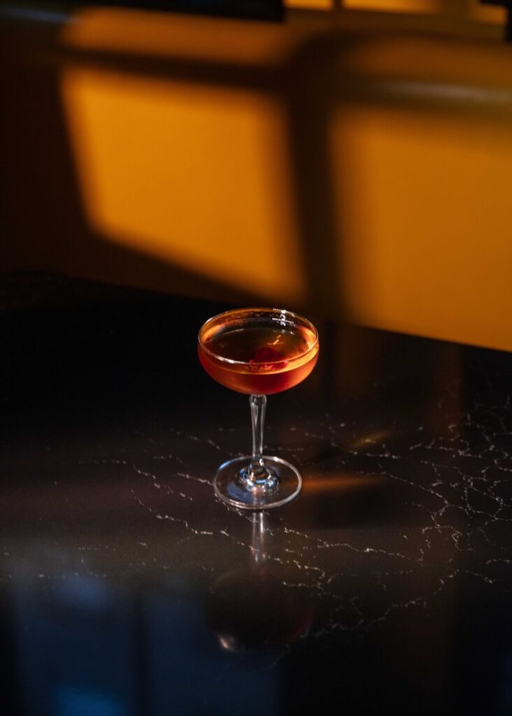 A cocktail in am amber color sits in a tall glass on a table in the evening light.