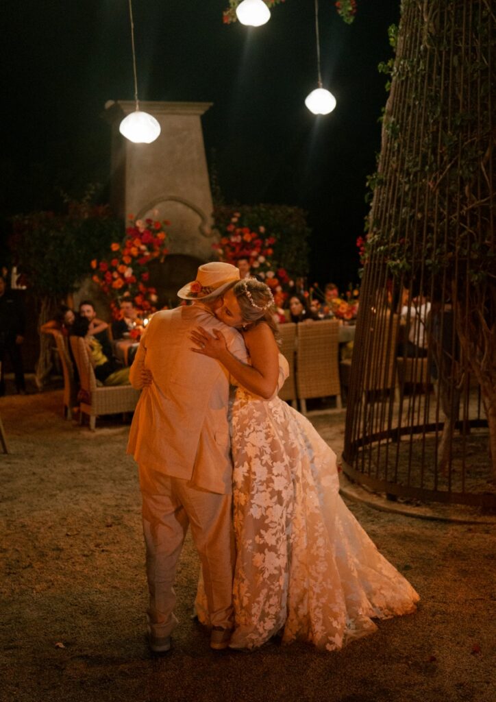 A bride in a floral lace gown hugging a guest in a tan suit and straw hat during an evening outdoor wedding reception at Villa Santa Cruz.