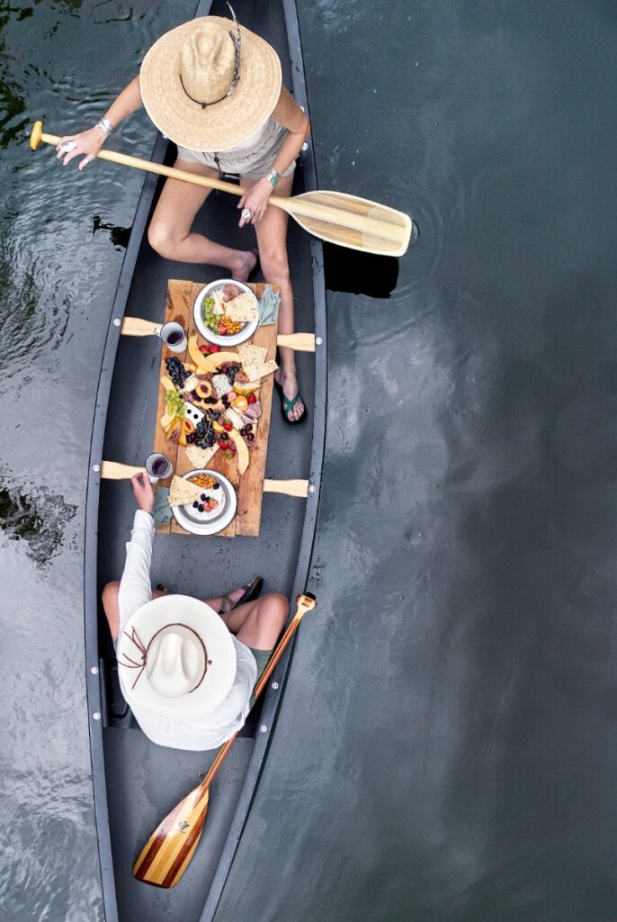 A couple in a canoe on a calm river, enjoying an elaborate charcuterie board lunch served on wooden planks across the boat's gunwales.