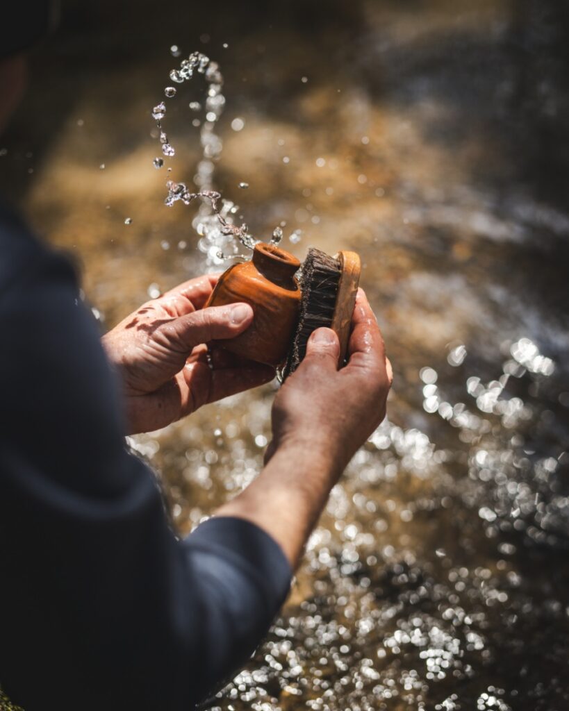 A person scrubbing a small, fired ceramic pot with a brush in a sparkling mountain stream to reveal its natural finish.