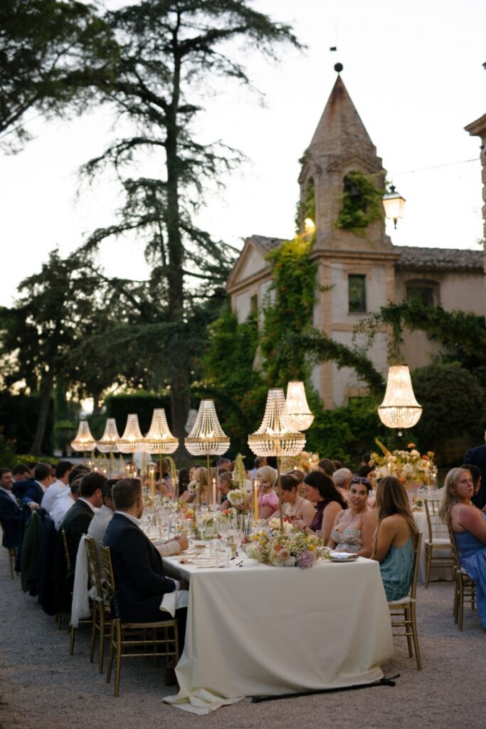 A sunset outdoor wedding reception in the garden of Relais Villa Corallo in Italy, featuring long tables with glittering chandeliers and floral centerpieces.