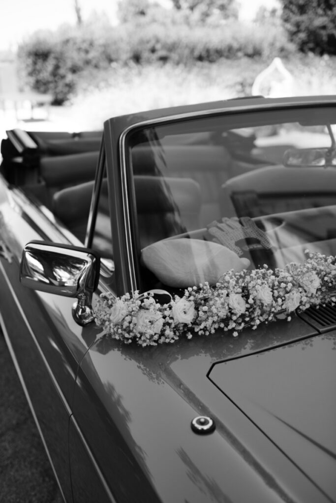 A black and white close-up of a vintage convertible wedding car adorned with a delicate white floral garland on the hood.