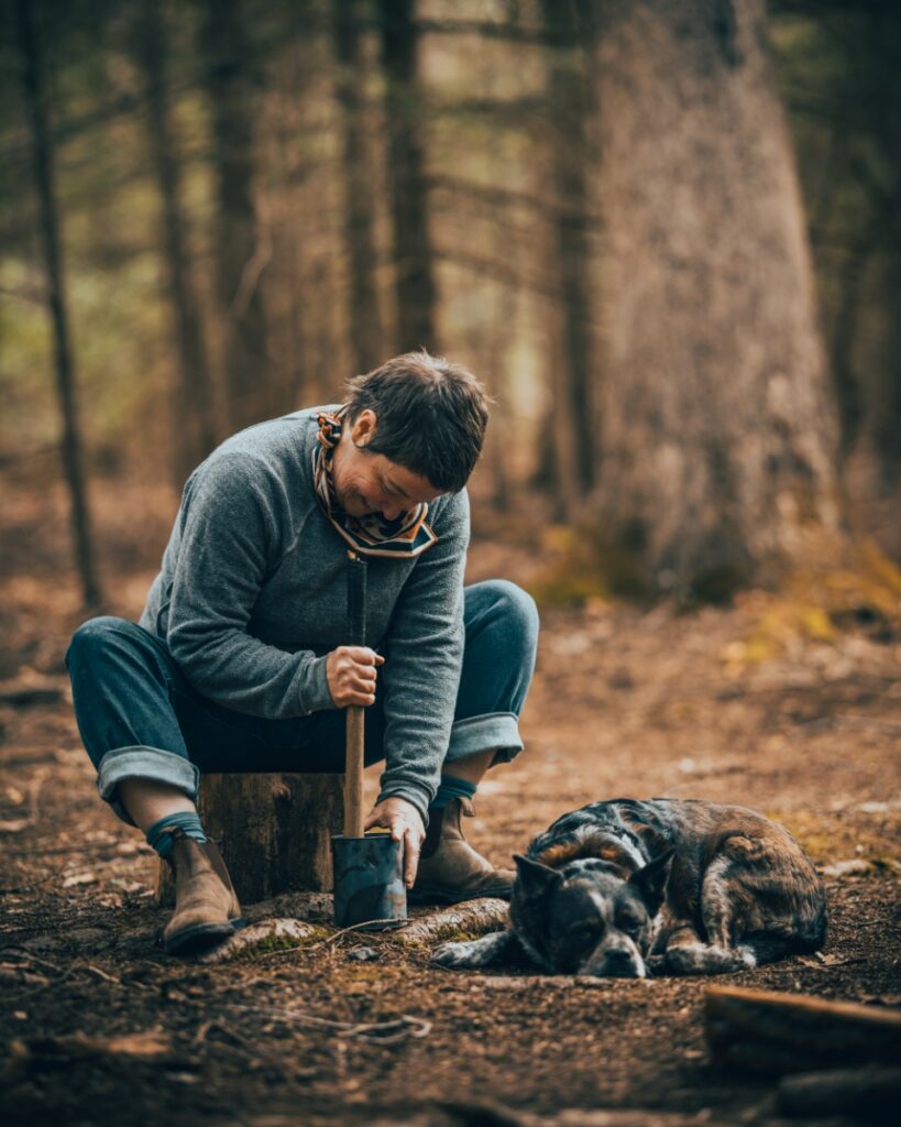 A woman smiling while using a wooden tool to pack or shape a ceramic pot in a forest setting, with a brindle dog resting nearby.