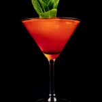 A martini glass with a red color cocktail and a mint garnish.