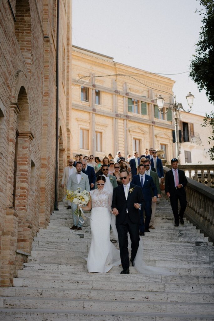 Bride Cristina Bartolacci and groom Philip Caputo leading their wedding guests down a stone staircase in the historic town of Colonnella, Italy.