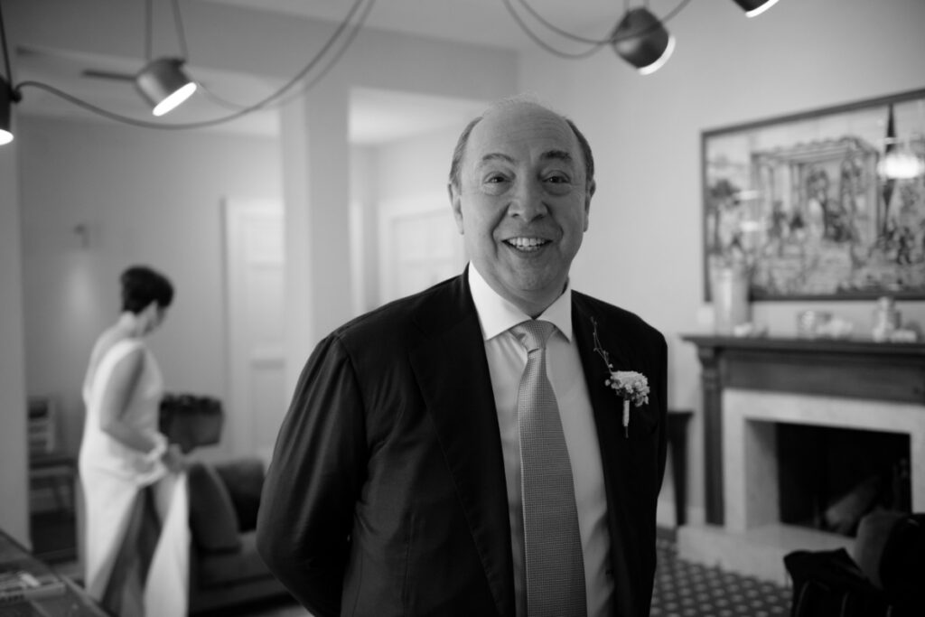 A black and white photo of Joseph Bartolacci, the bride's father, smiling in a suit and tie before the wedding ceremony.