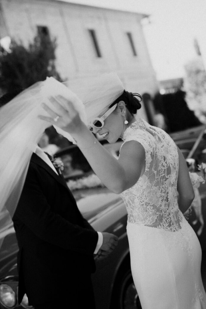 Bride Cristina Bartolacci in a lace Ines de Santo wedding gown and white sunglasses, lifting her veil during her wedding in Italy.