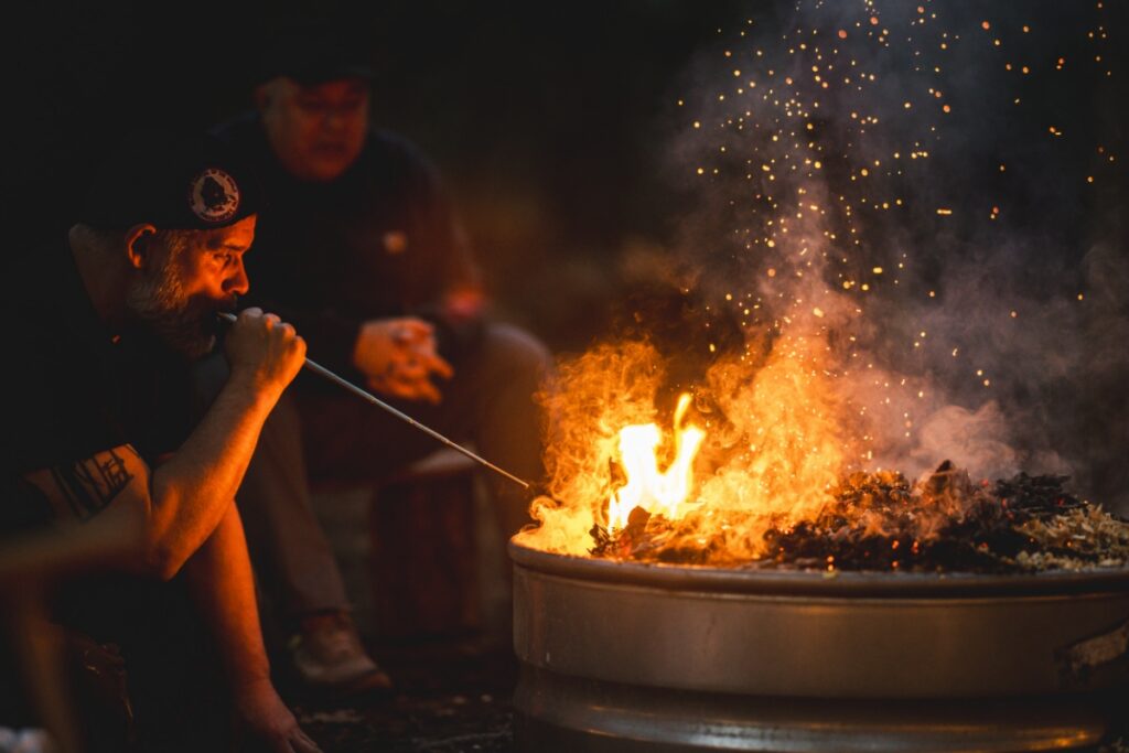 A man using a long metal tube to blow air into a roaring campfire to increase the heat for a primitive pottery firing in the woods at night.