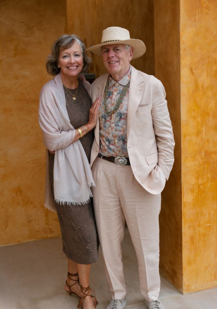 A smiling wedding guest in a brown dress and wrap standing next to a man in a tan suit, floral shirt, and straw hat at Villa Santa Cruz.